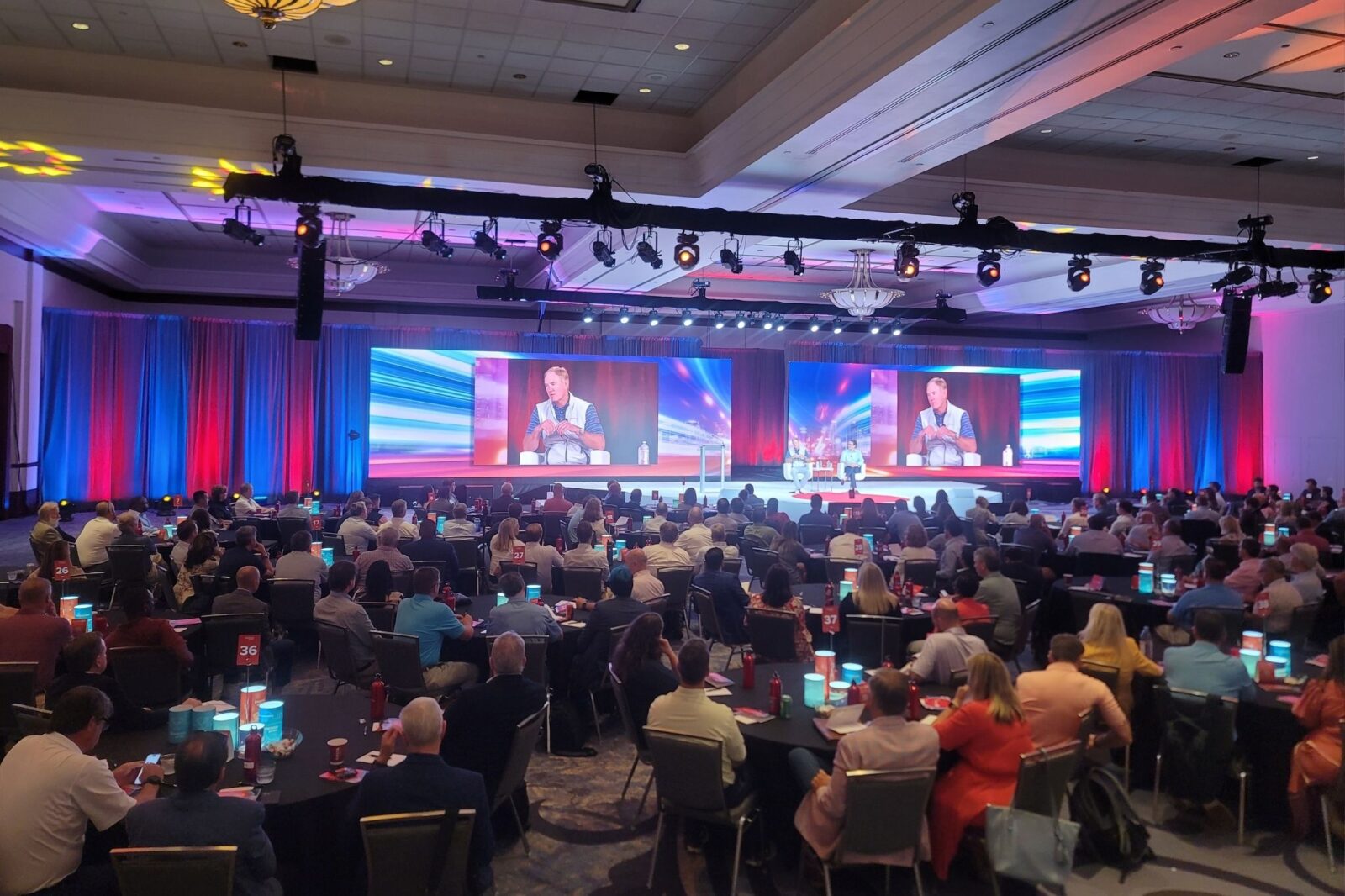 Large corporate conference ballroom with full audience seating, dual LED video walls, stage lighting truss, and live panel discussion produced by Innovent Technologies