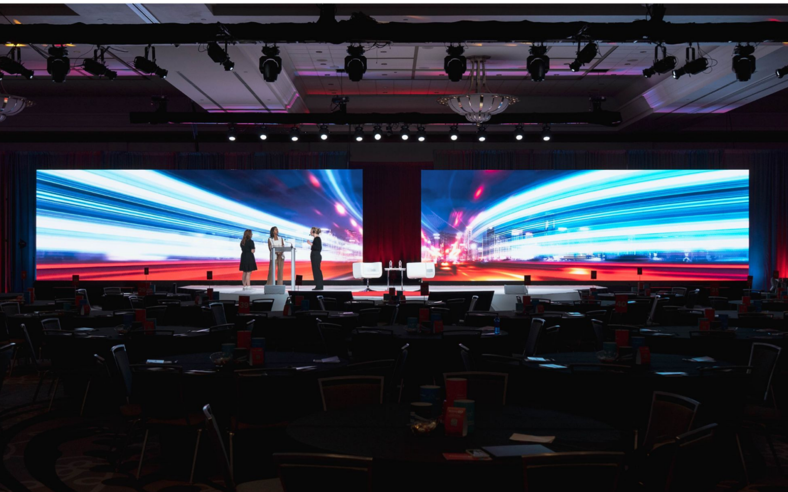 Corporate general session stage with dual LED video walls, presenters at podium, white lounge seating, and ballroom round table setup produced by Innovent Technologies