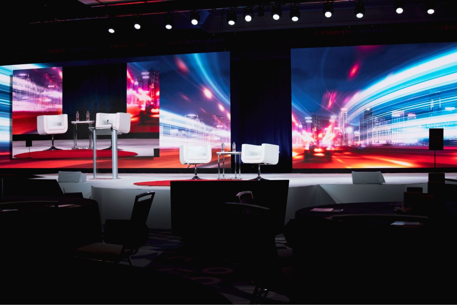 Corporate event stage with dual LED video walls, white lounge chairs, podium, and red circular accent carpet produced by Innovent Technologies