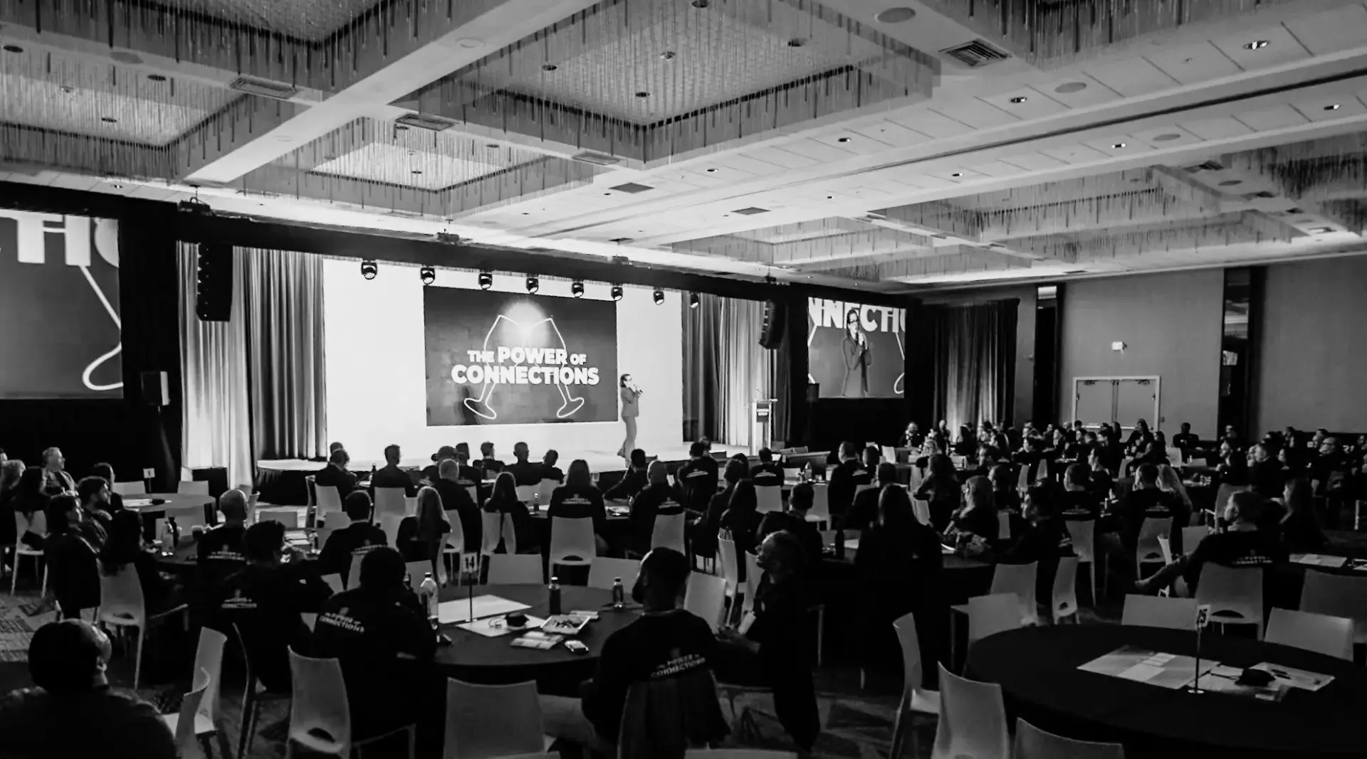 Corporate conference general session with stage presentation, large projection screen, and audience seated at round tables during multi-day event
