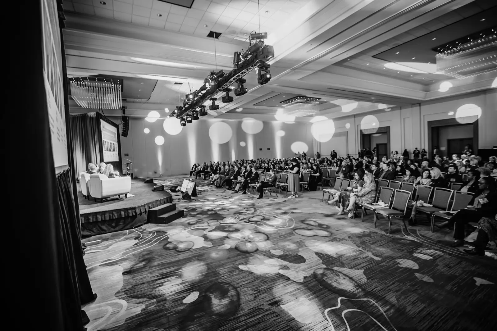 Conference general session with stage lighting, production rigging, speakers, and an engaged audience.