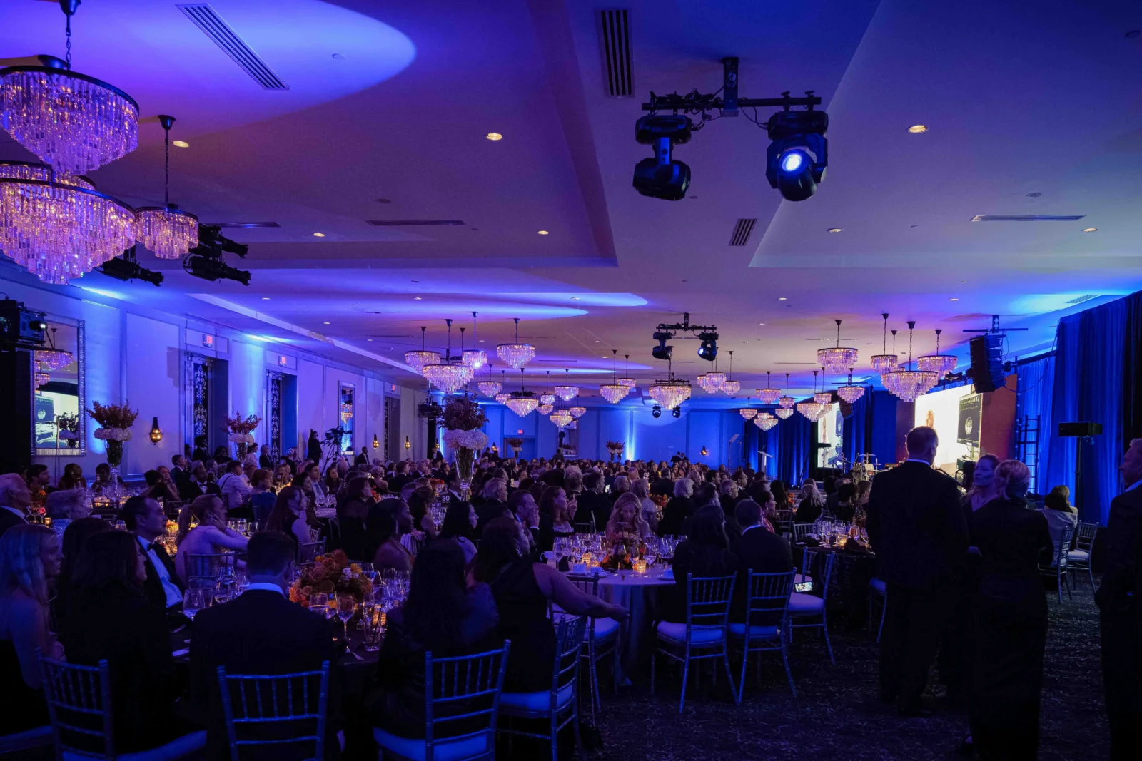 Large gala audience seated at banquet tables in a ballroom illuminated by blue and purple event lighting, with presentation screens and staging.